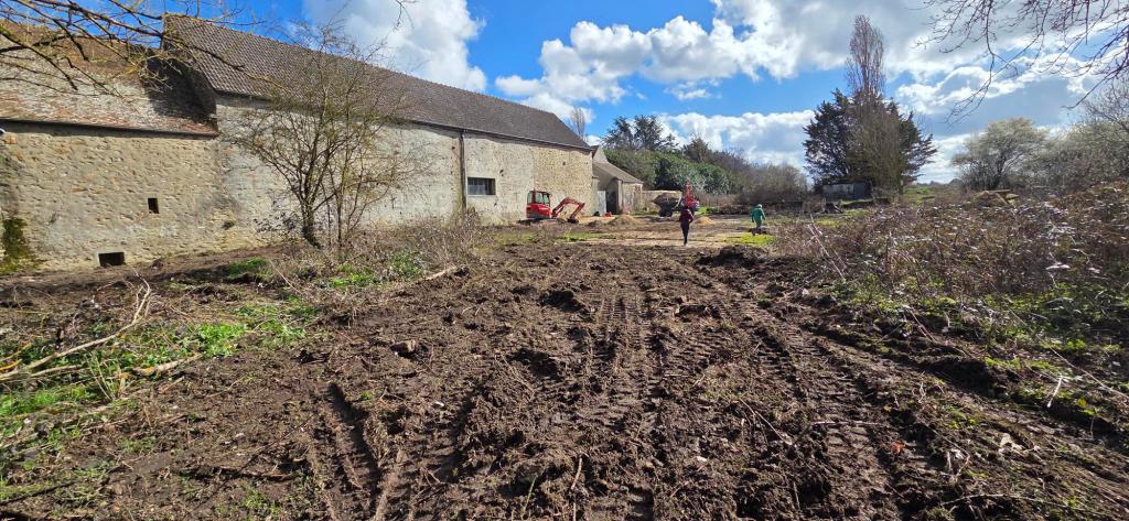Vue d'une cour de ferme avec des traces de pneus, des bâtiments en pierre et un ciel nuageux.