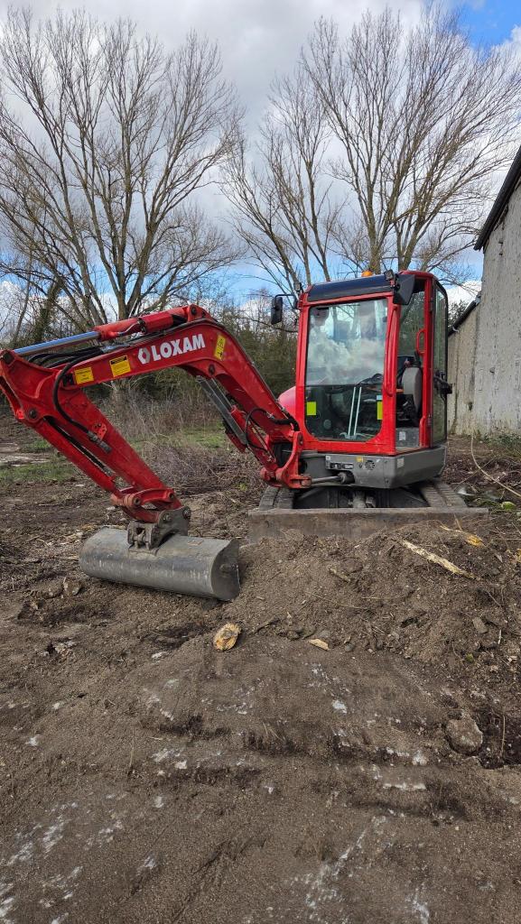 Une excavatrice rouge sur un terrain avec des arbres et un ciel nuageux en arrière-plan.